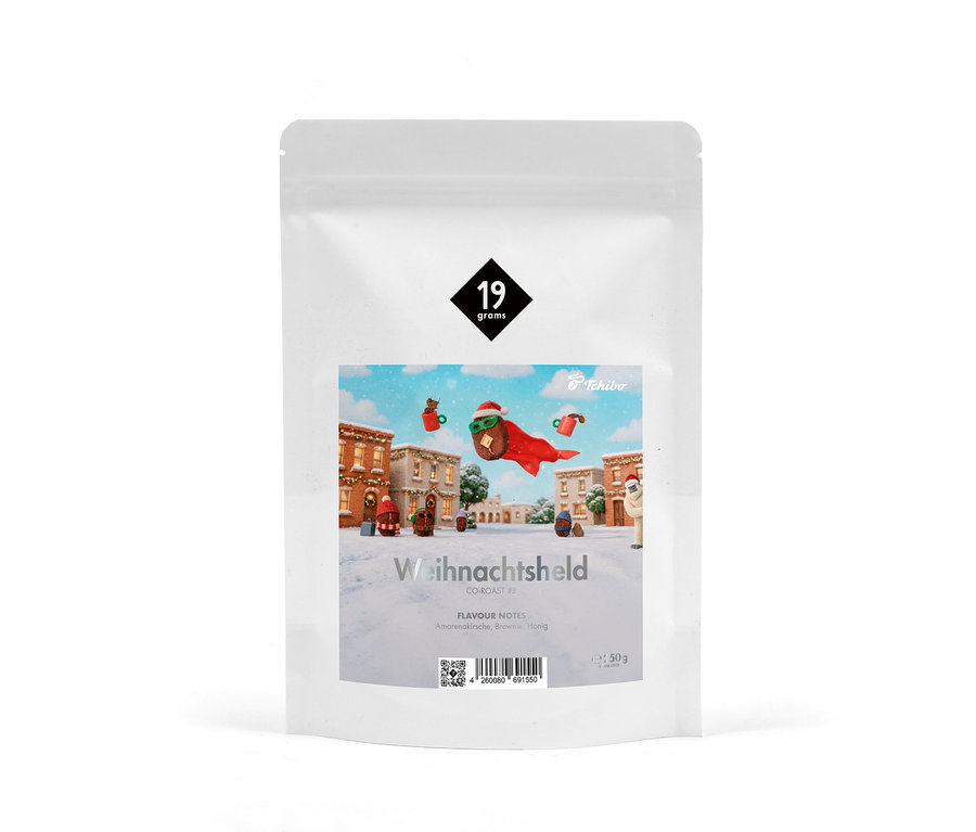 Eine weiße Kaffeeverpackung mit einem Etikett, das ein verschneites Weihnachtsdorf und einen fliegenden Kaffeehelden mit Weihnachtsmütze und rotem Umhang zeigt. Text: „Weihnachtsheld“, „19 grams“, „Tchibo“, „Amarenakirsche, Brownie, Honig“.