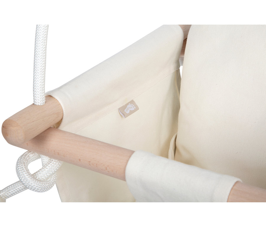 Nahaufnahme der small foot Babyschaukel Komfort mit cremefarbenem Stoff, hellen Holzstangen und weißen Seilen; ein kleines Etikett mit Fußabdruck-Logo ist sichtbar.