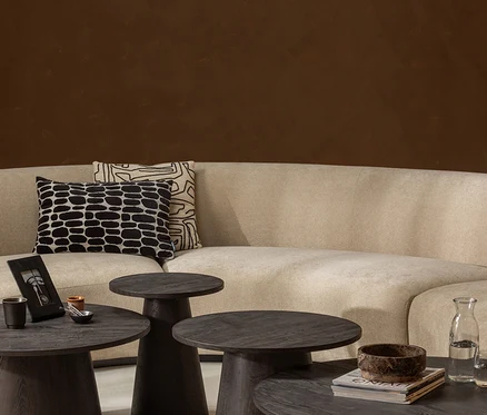 Beiges Sofa mit zwei gemusterten Kissen vor einer braunen Wand, davor vier runde Couchtische aus dunklem Holz. Auf den Tischen stehen ein Bilderrahmen, eine Tasse, eine Schale, Bücher, eine Schüssel und eine Wasserflasche.