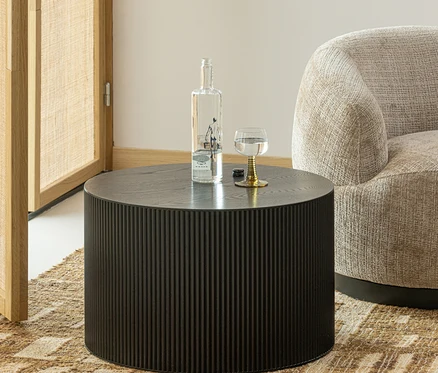 Runder, dunkler Couchtisch mit gerippter Seite, darauf eine klare Flasche und ein Glas mit goldenem Stiel, neben einem hellen Sessel auf einem gemusterten Teppich.
