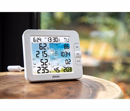 Weisse digitale Wetterstation von Alecto auf einem Holztisch, die Innen- und Aussentemperatur, Luftfeuchtigkeit, Wind, Luftdruck, Regen, UV und Licht anzeigt.