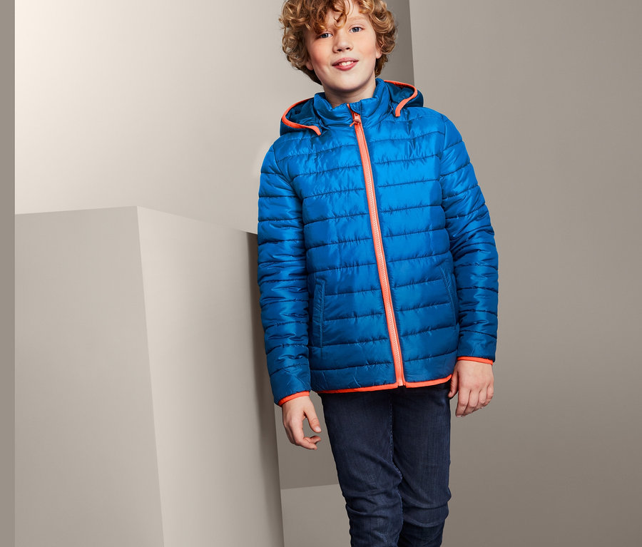 Junge in blau gesteppter Jacke mit Farbverlauf, dunklen Jeans und Turnschuhen steht an eine Wand gelehnt.