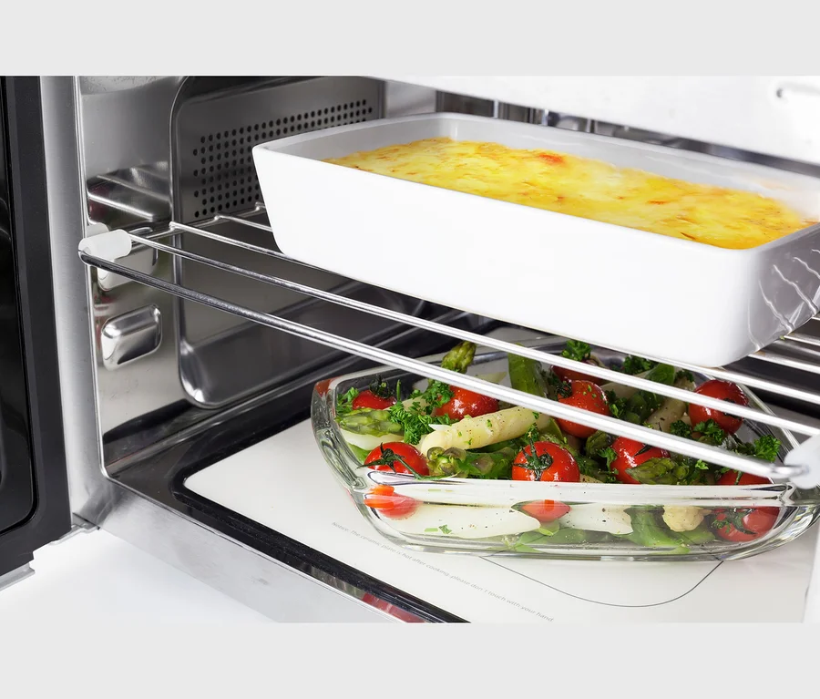 Zwei Gerichte garen in einem Ofen: eine weiße, rechteckige Form mit gelbem Gratin auf dem oberen Rost und eine Glasschale mit Spargel, Kirschtomaten und Kräutern auf dem unteren Rost.