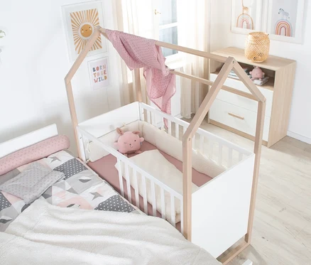 Weißes und holzfarbenes Hausbettchen mit rosa Bettwäsche und Hasenplüschtier, daneben ein Bett mit gemusterter Bettwäsche. Im Hintergrund eine Kommode mit Deko und Wandbilder.