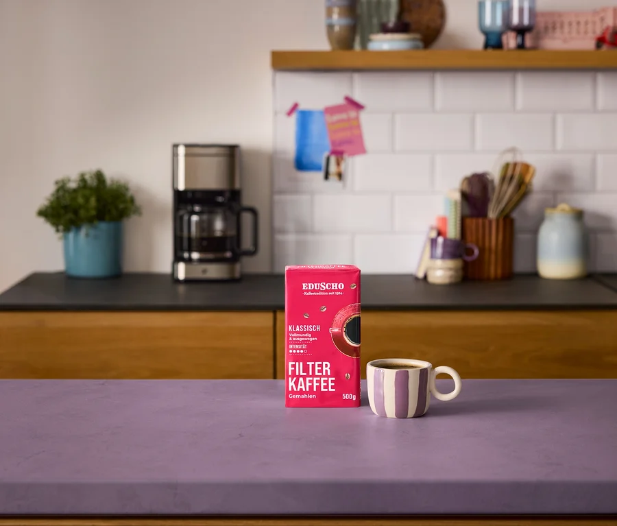 Eine rote Packung »Eduscho Filterkaffee Klassisch - 500 g Gemahlen« und eine gestreifte Kaffeetasse stehen auf einer lila Arbeitsplatte. Im Hintergrund ist eine silberne »Let's Brew Elegant« Filterkaffeemaschine auf einer schwarzen Arbeitsplatte zu sehen.