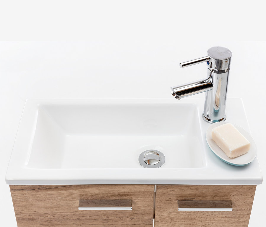 Weißes Keramikwaschbecken mit Holzschrank, Chromarmatur und Seife auf Unterlage.