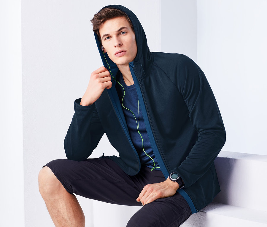 Mann sitzt auf einer weißen Oberfläche, trägt eine dunkelblaue Sportjacke mit Kapuze, schwarze Shorts und eine Uhr.