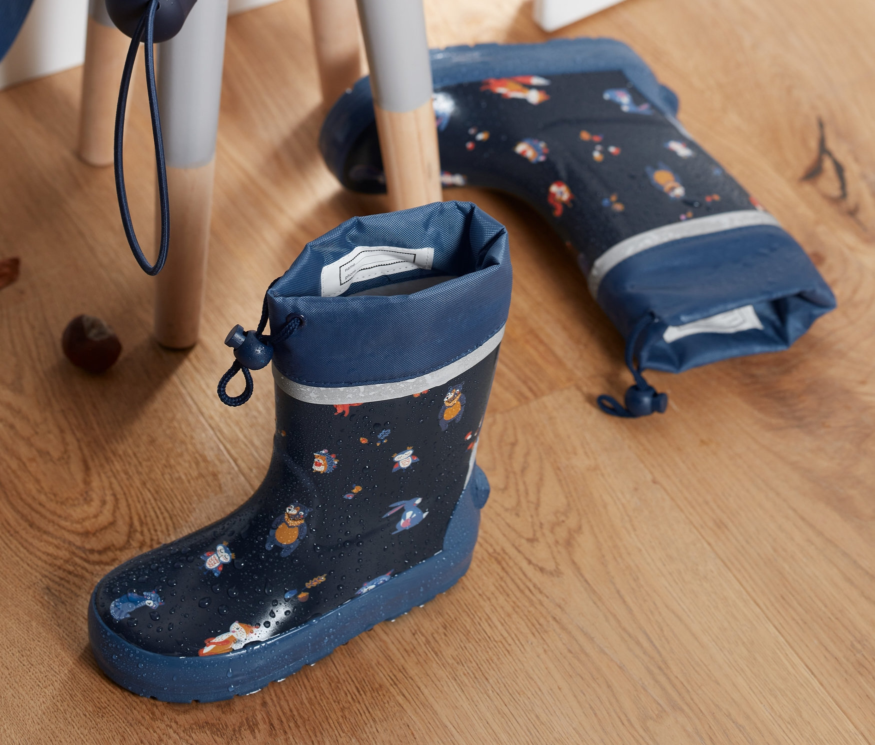 Dunkelblaue Gummistiefel mit Alloverprint stehen nebeneinander auf einem Holzboden, ein zusammengefalteter Regenschirm und Kastanien auf einem Hocker.