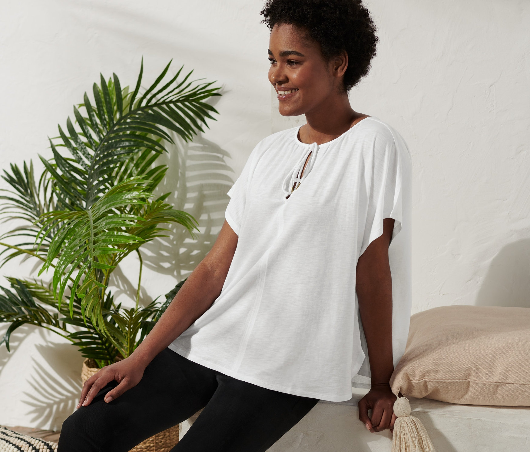 Eine Frau sitzt und lächelt, sie trägt ein Blusenshirt mit Bindeband, schwarze Hosen und Plateausandalen.