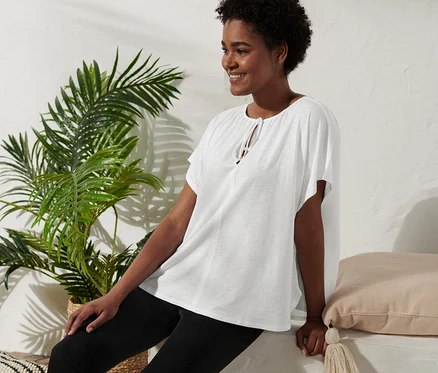 Eine Frau sitzt und lächelt, sie trägt ein Blusenshirt mit Bindeband, schwarze Hosen und Plateausandalen.