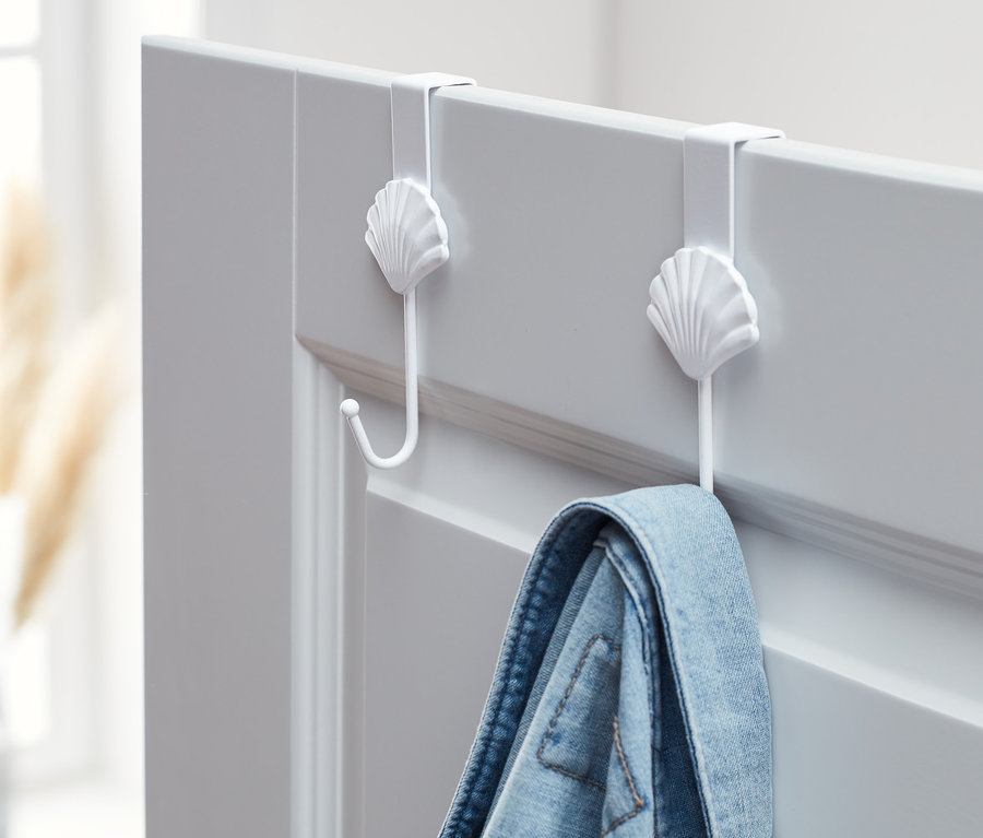 Zwei weiße Türhaken im Muscheldesign halten eine blaue Jeansjacke an einer grauen Tür.