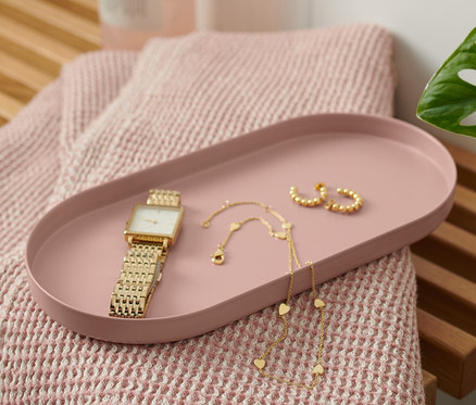 Auf einer Holzbank liegen ein rostfarbenes Tablett aus Metall, eine Uhr, eine Halskette und Ohrringe. Neben dem Tablett liegen ein rosa Handtuch und eine Flasche mit Spender.