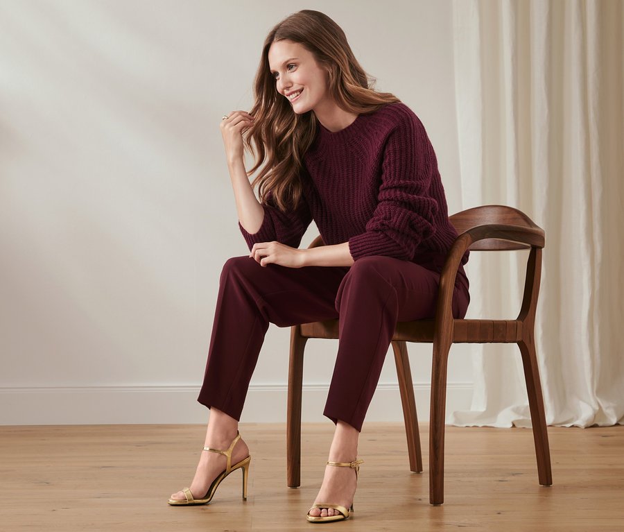 Frau sitzt auf einem Stuhl und trägt eine Stoffhose und einen Pullover.