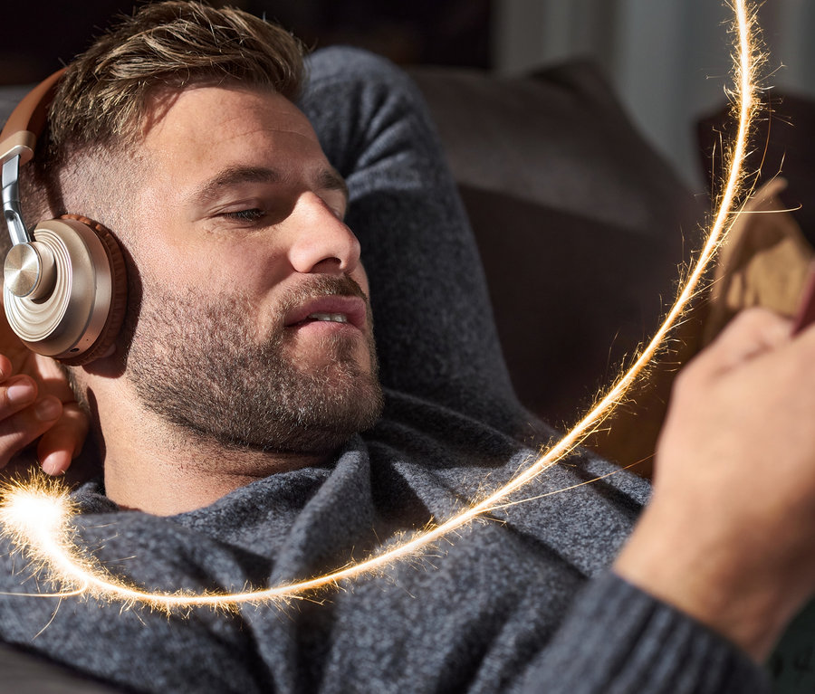 Mann liegt auf einer Couch und trägt On-Ear-Bluetooth®-Kopfhörer und schaut auf sein Handy.