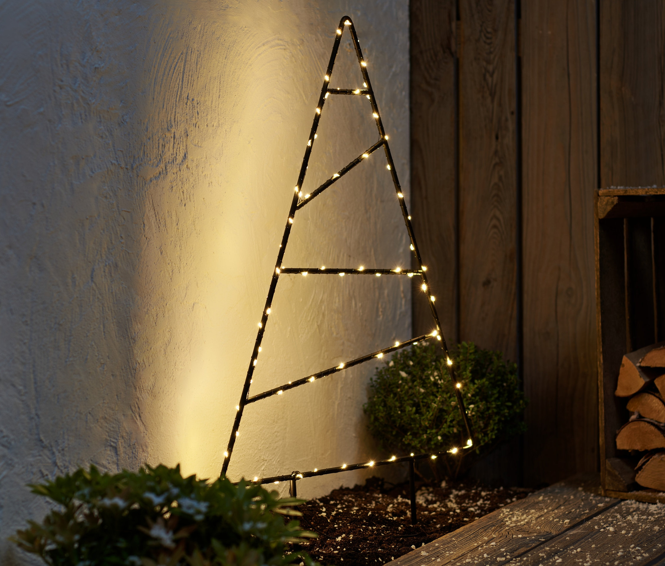Eine große LED-Outdoor-Tanne aus Metall steht neben einer Holzwand.