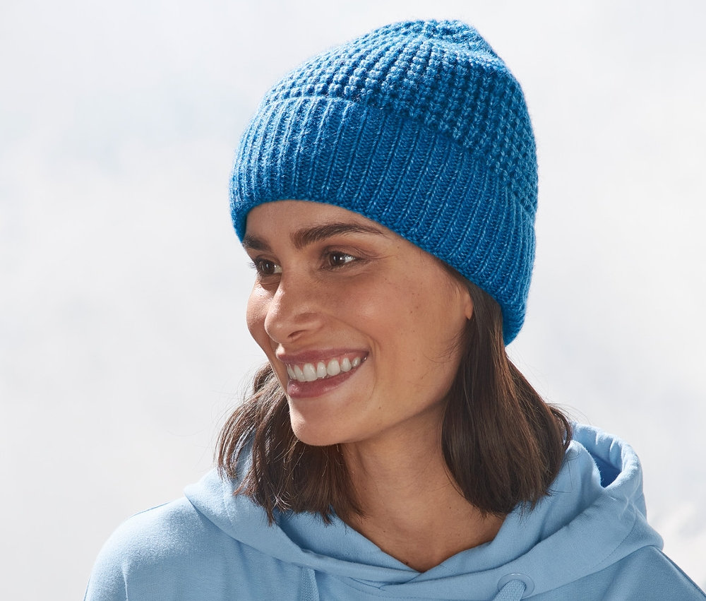 Frau mit verschränkten Armen vor Bergkulisse trägt eine blaue Strickmütze.