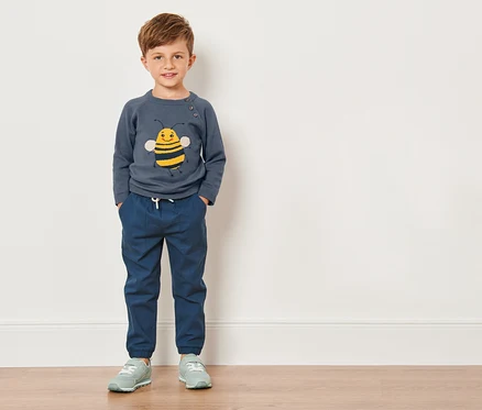 Ein Junge in einem Kleinkinder-Feinstrickpullover mit Bienenmotiv steht vor einer weißen Wand.