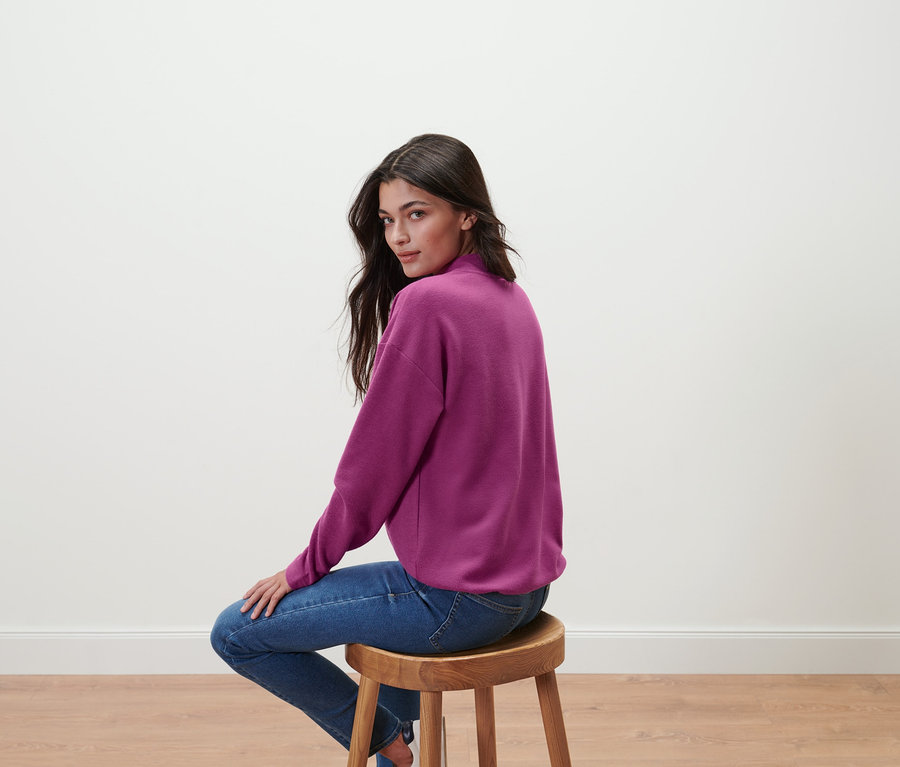 Frau sitzt auf einem Hocker und trägt ein lila Sweatshirt mit Stehkragen und Jeans.