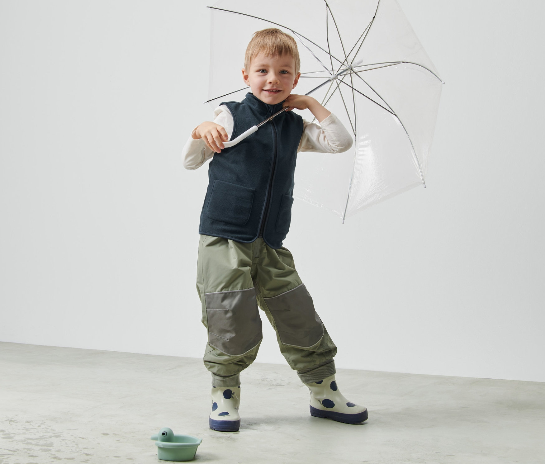 Junge hält einen transparenten Regenschirm und trägt eine dunkelblaue Kinder-Fleeceweste.