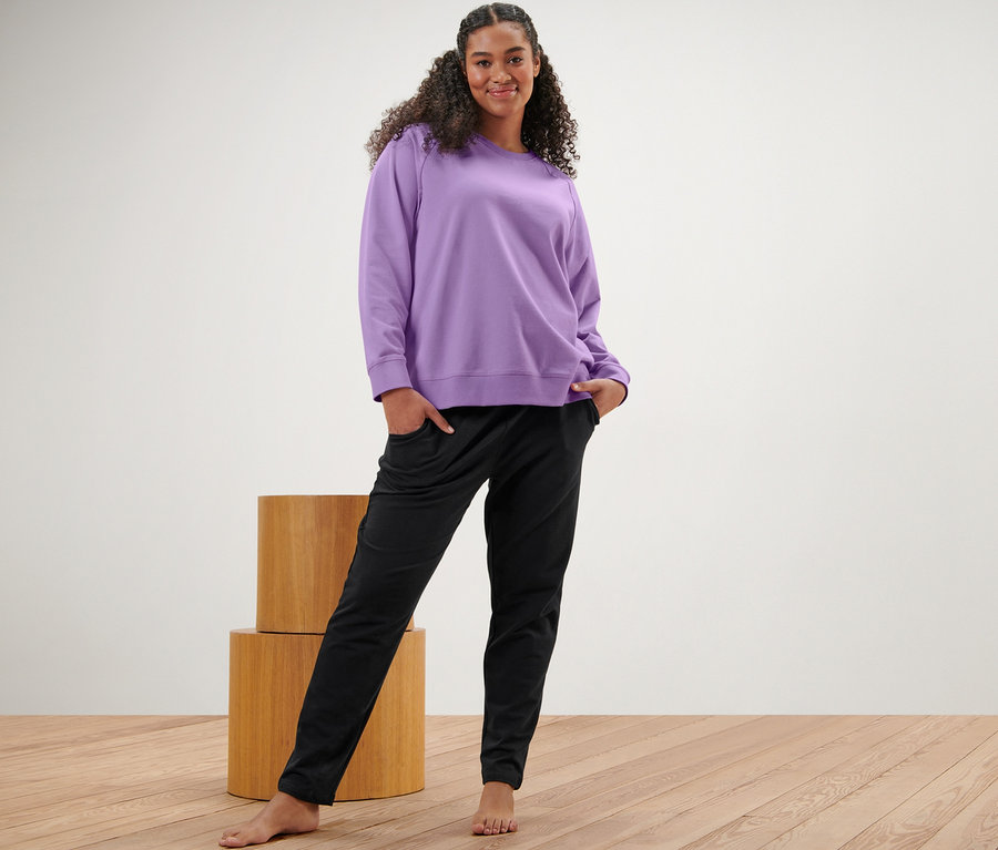 Frau steht mit Händen in den Taschen einer schwarzen Sweathose und einem fliederfarbenen Yogasweatshirt.