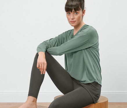 Frau sitzt auf einem Holzhocker und trägt ein 2-in-1-Sportshirt in Salbei und Leggings.