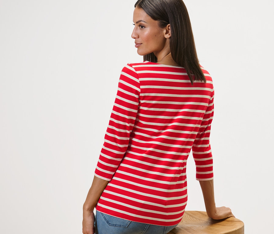 Frau sitzt seitlich auf einem Holzhocker und trägt ein gestreiftes Shirt mit 3/4-Arm, rot getreift.