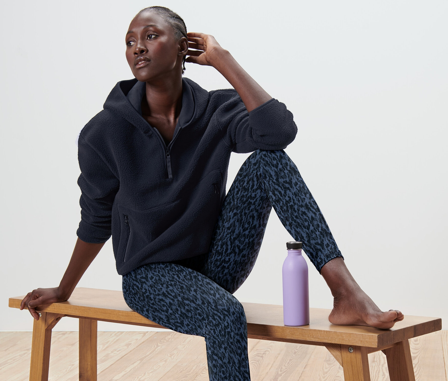 Frau sitzt mit Teddy-Kapuzenpullover und gemusterten Leggings auf einer Bank, daneben eine lila Flasche.
