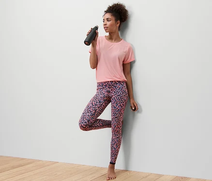 Frau im rosa T-Shirt balanciert auf einem Bein und trinkt aus einer schwarzen Flasche. Sie trägt eine Wende-Sporttight, rosa/navy.