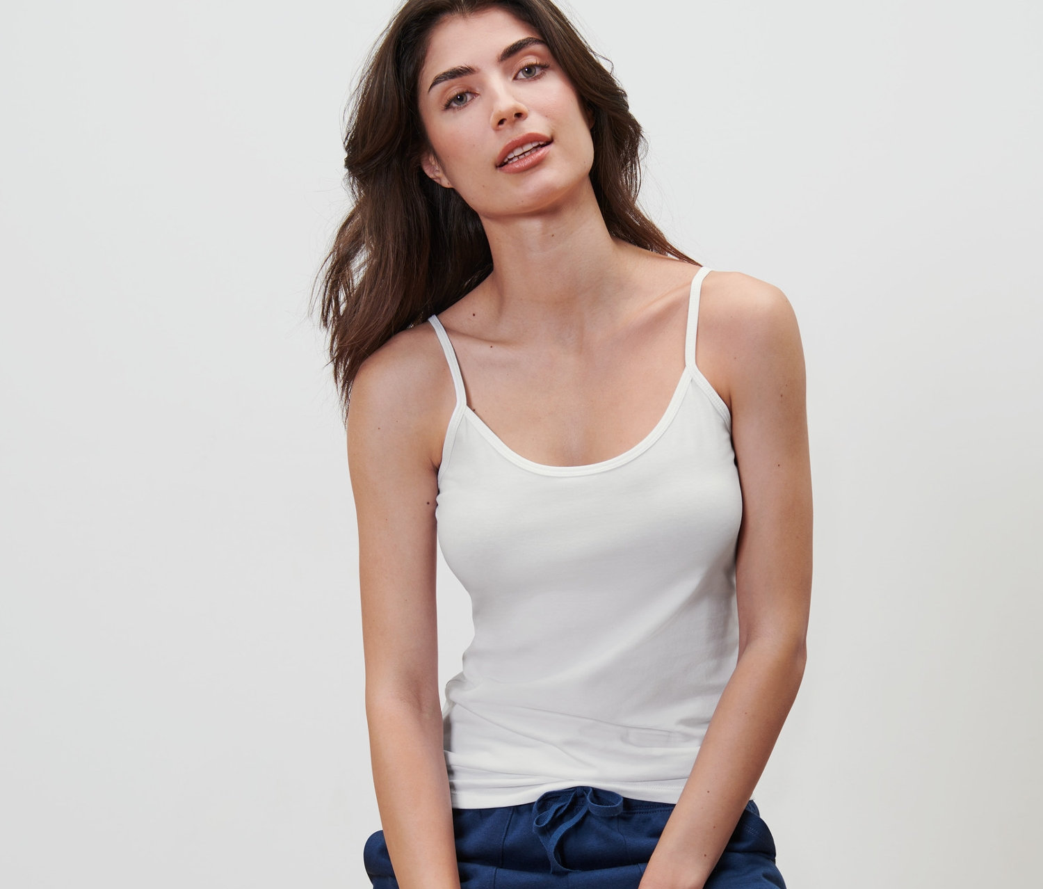 Frau sitzt auf einem Hocker und trägt ein weißes Spaghetti-Hemdchen aus einem 5er-Pack.