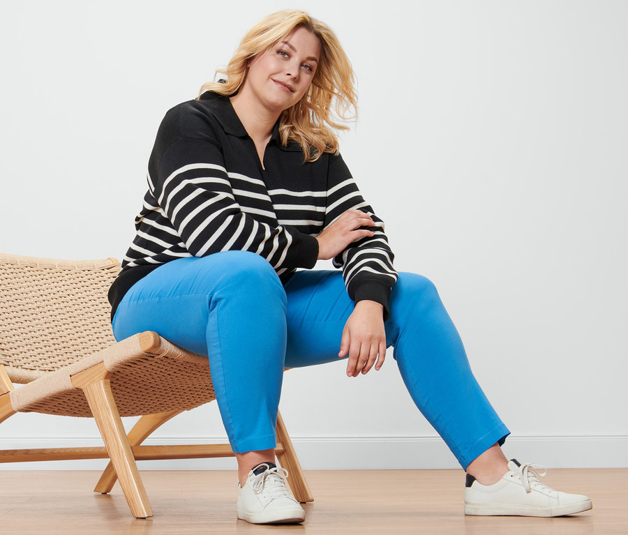 Eine blonde Frau sitzt auf einem Korbsessel und trägt einen Pullover mit Kragen, eine Bengalin-Hose und Ledersneaker.