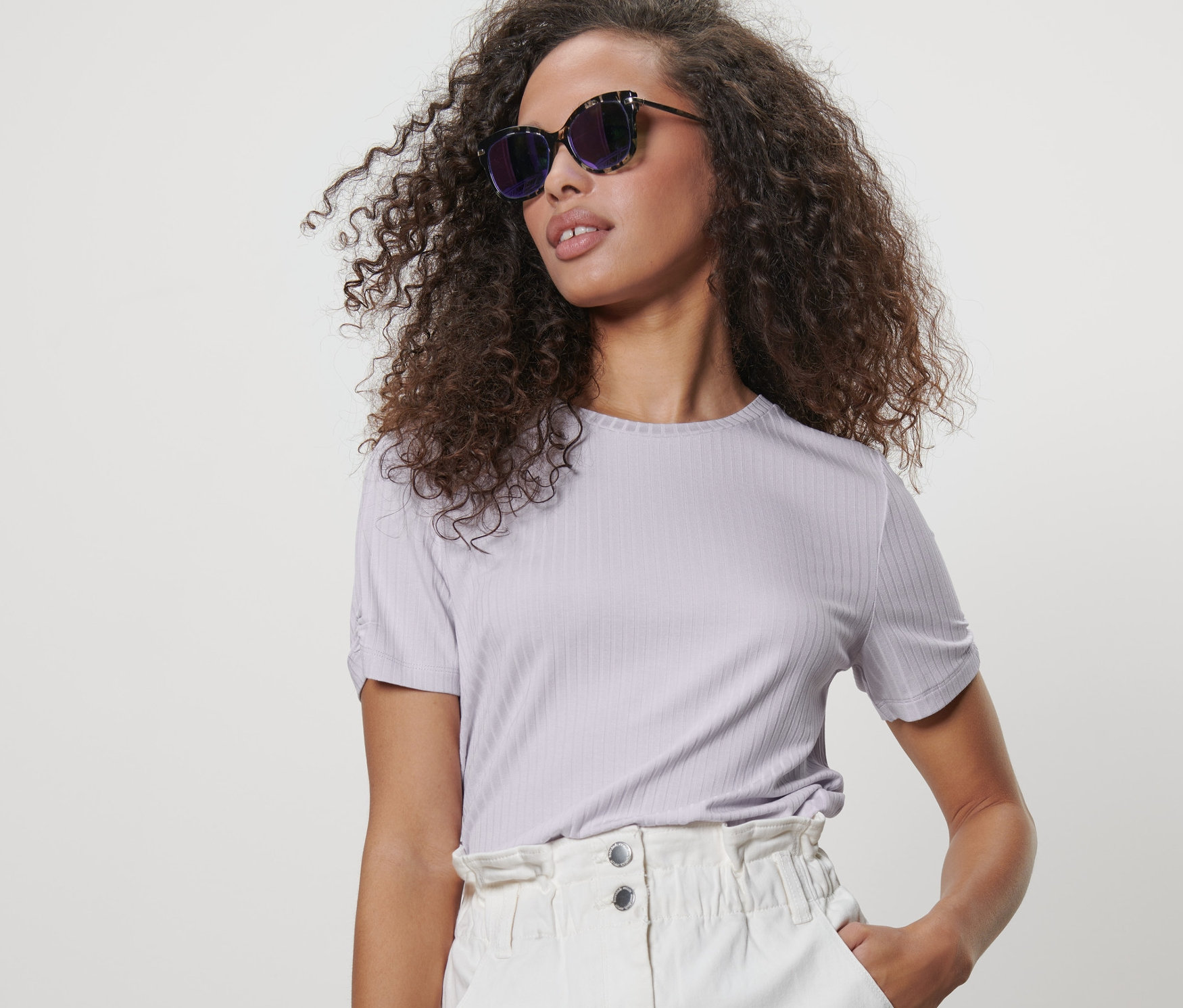 Frau mit braunen lockigen Haaren posiert in einem fliederfarbenen, gerippten Shirt und einem weißen Rock.