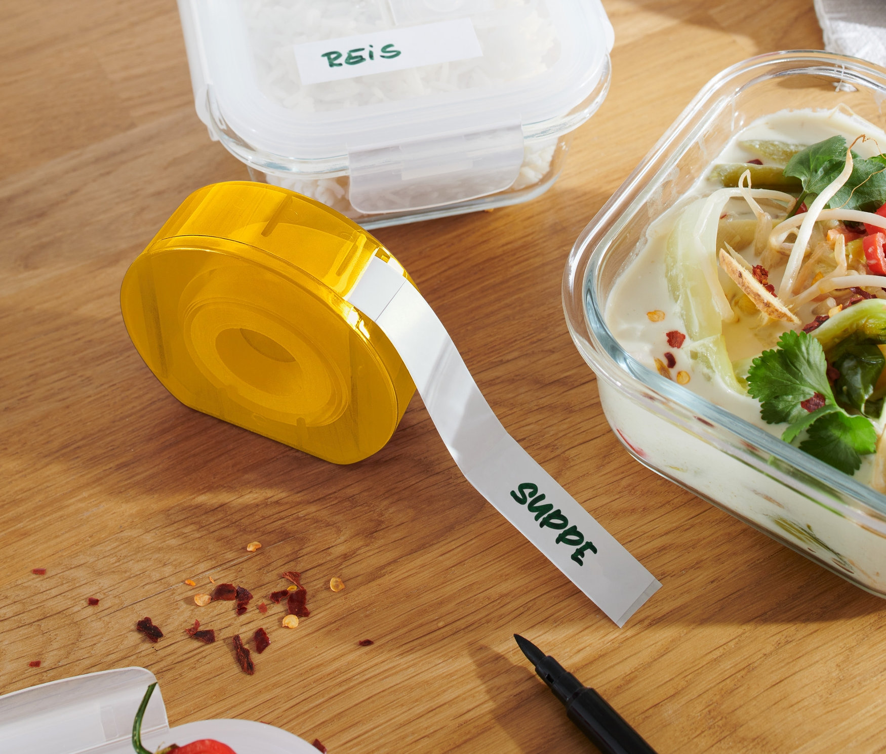 Gelber Etikettenspender auf Holztisch neben Behältern mit Reis und Suppe.