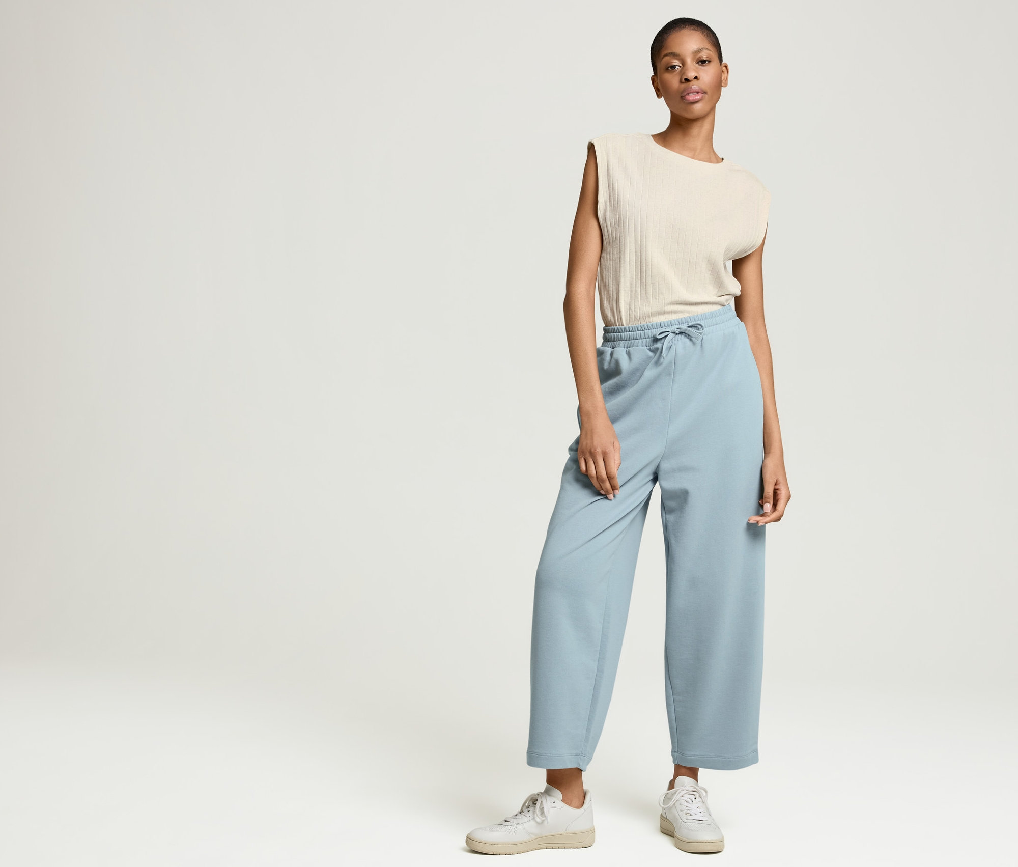 Frau posiert im Studio in hellblauer NAH/STUDIO Joggpants aus Bio-Baumwolle und einem gerippten NAH/STUDIO Shirt aus ungefärbtem Baumwoll-Hanf-Mix.