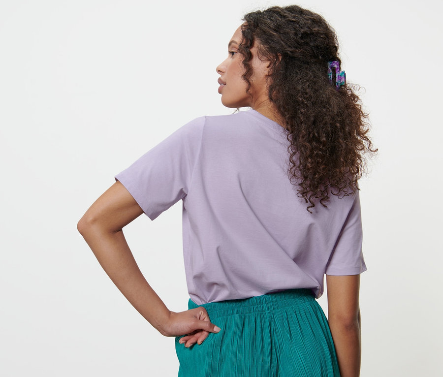 Frau mit braunen Haaren trägt ein lila T-Shirt mit Stickerei und eine grüne Hose.