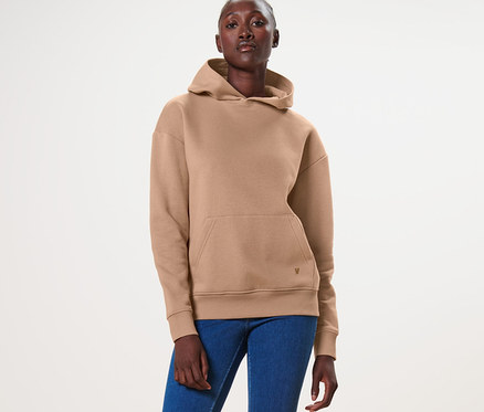 Frau steht und trägt einen beigen Hoodie, blaue Jeans und weiße Turnschuhe.