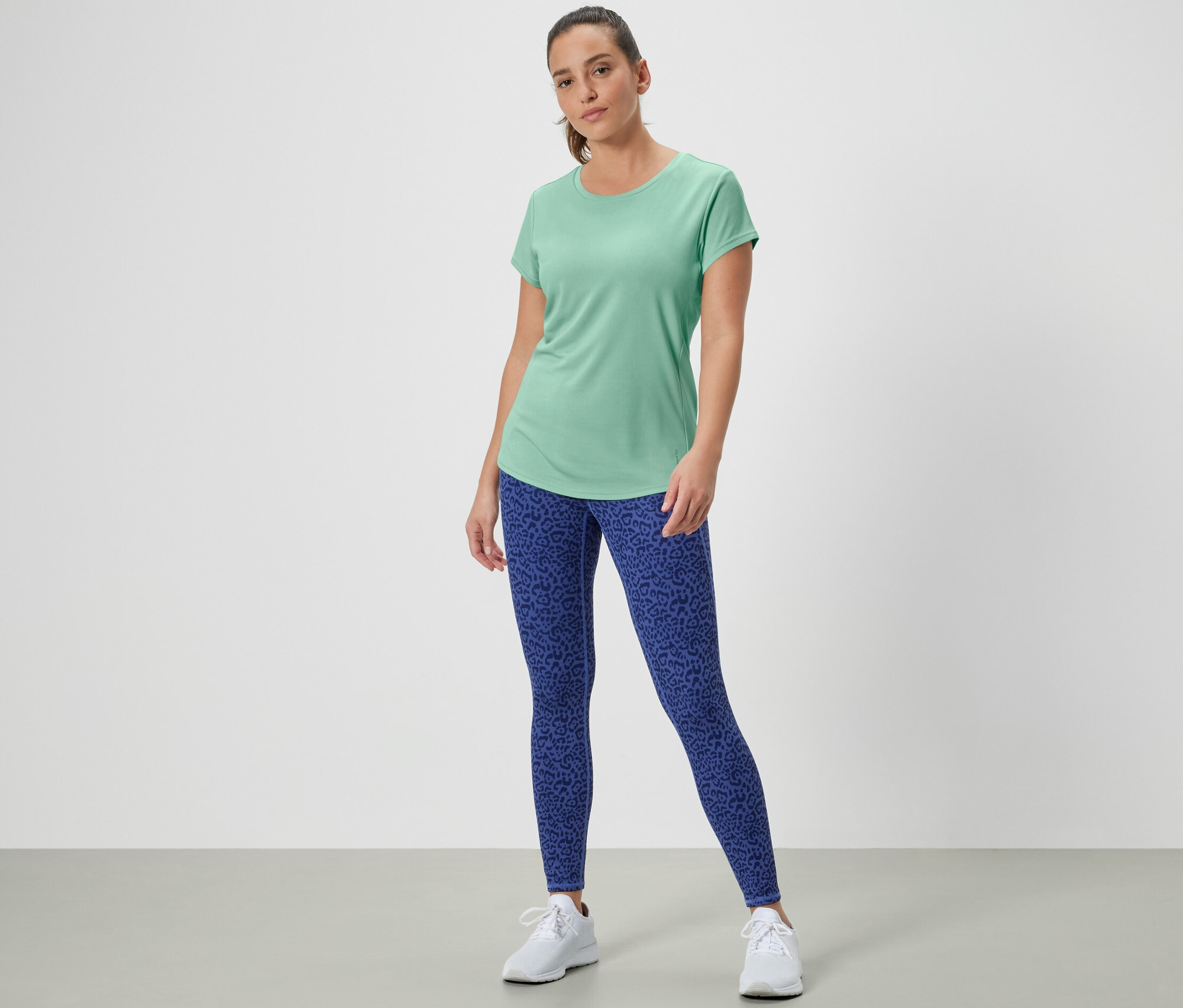 Frau steht in grünem Sportshirt und blauen Leggings.
