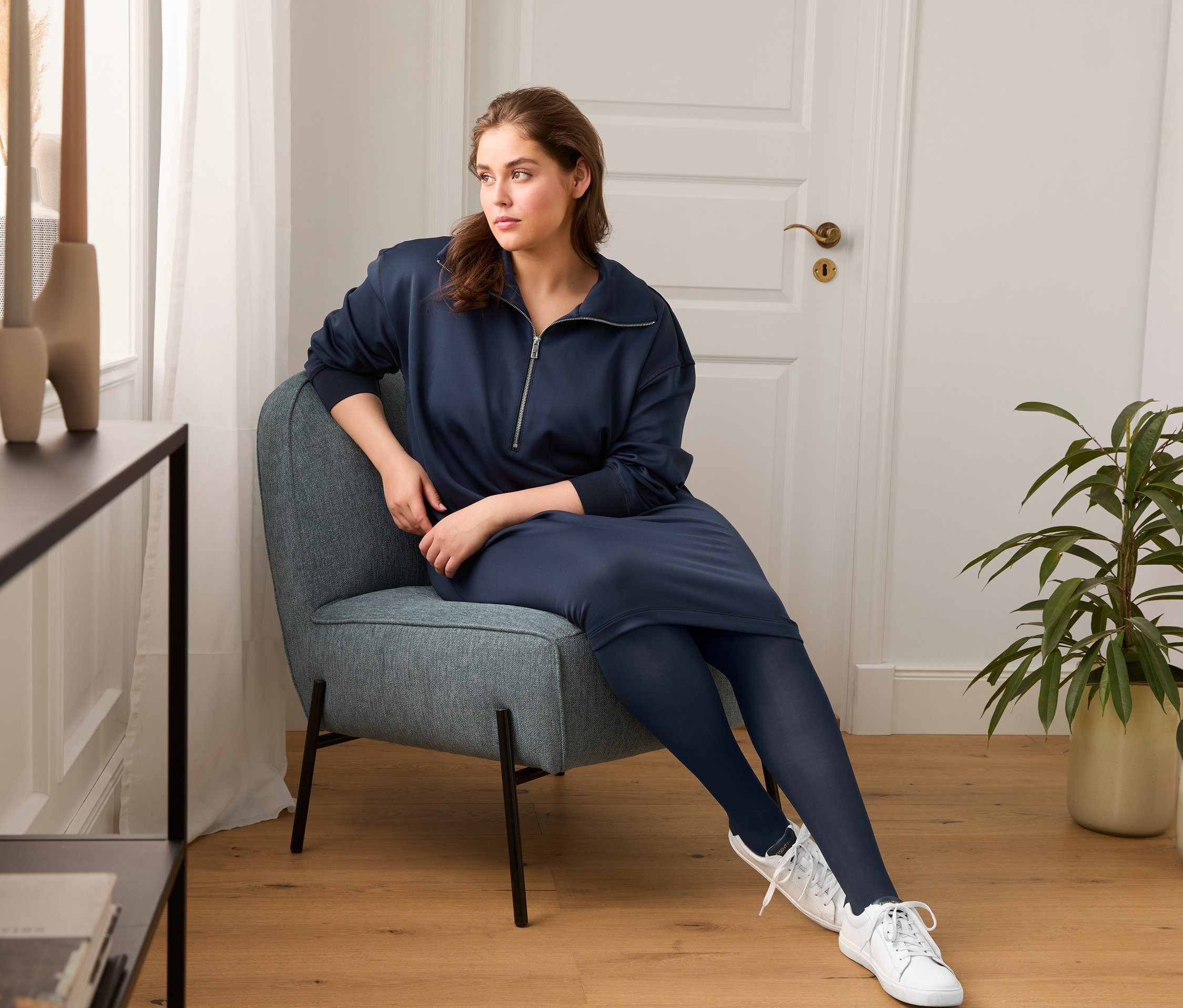 Eine Frau sitzt in einem Sessel und trägt ein Sweatkleid, eine blickdichte Strumpfhose und Sneaker aus chromfrei gegerbtem Leder.