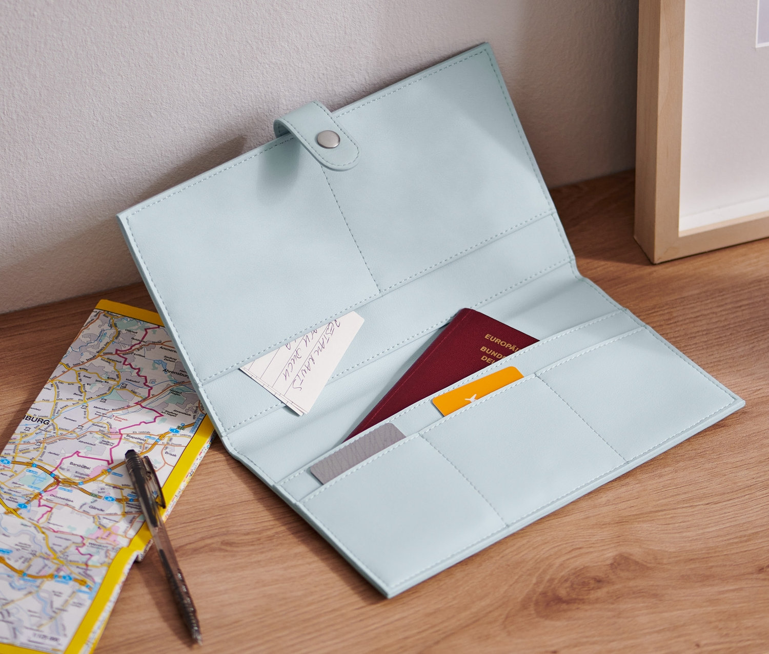 Ein offener, hellblauer Reise-Organizer liegt auf einem Holztisch neben einer Landkarte und einem Stift.