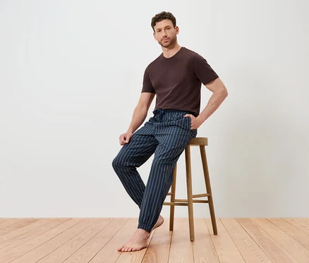 Mann sitzt auf einem Holzhocker, trägt ein braunes T-Shirt und eine blau gestreifte Hose.