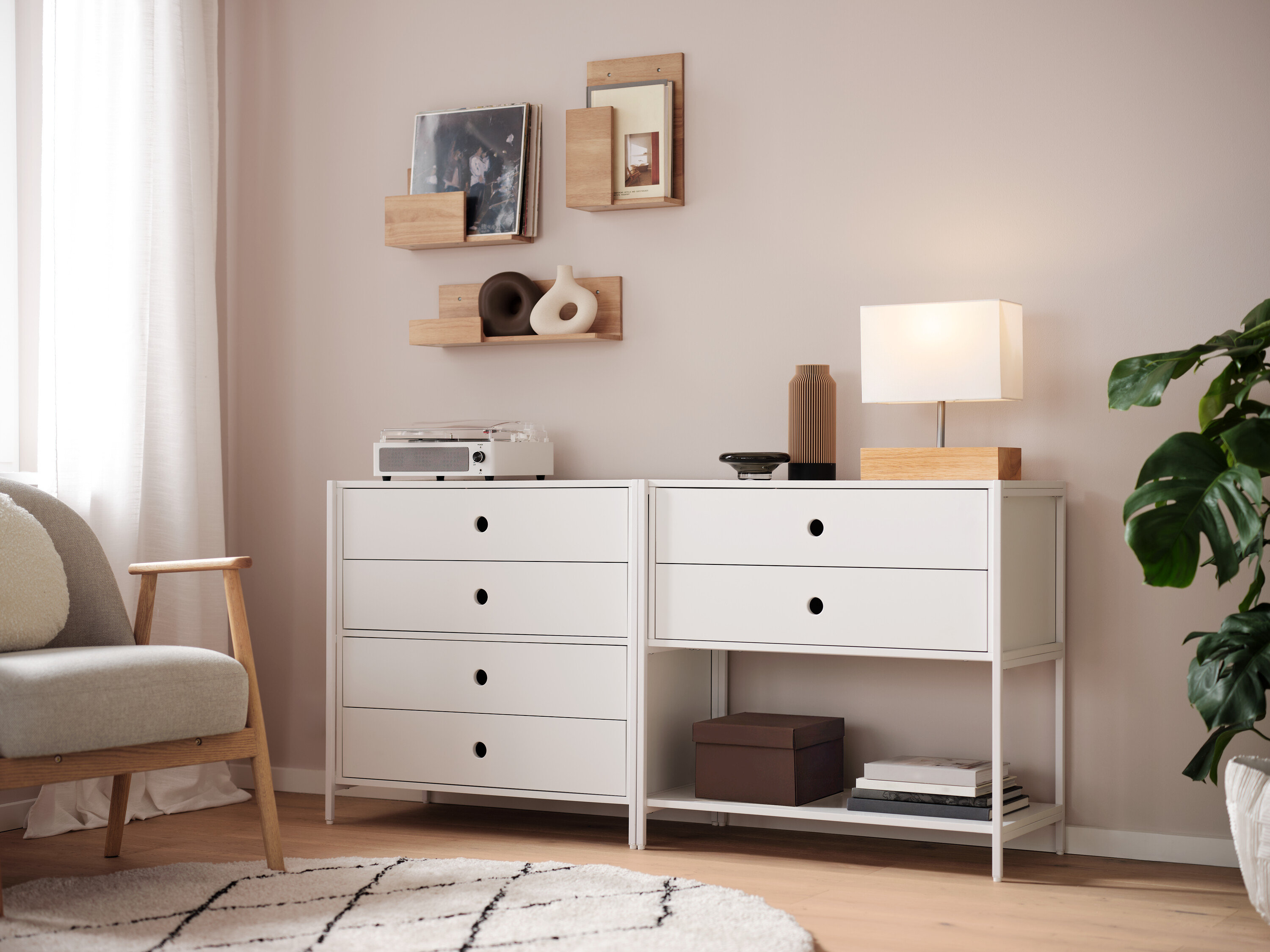 Zwei weiße Kommoden stehen nebeneinander, darauf eine Lampe, eine Vase und ein Radio. An der Wand hängen Regale.