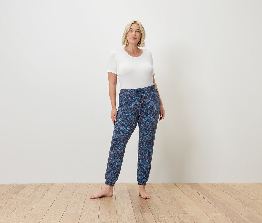 Frau steht barfuß auf einem Holzboden und trägt ein weißes T-Shirt und blaue Jogginghosen.