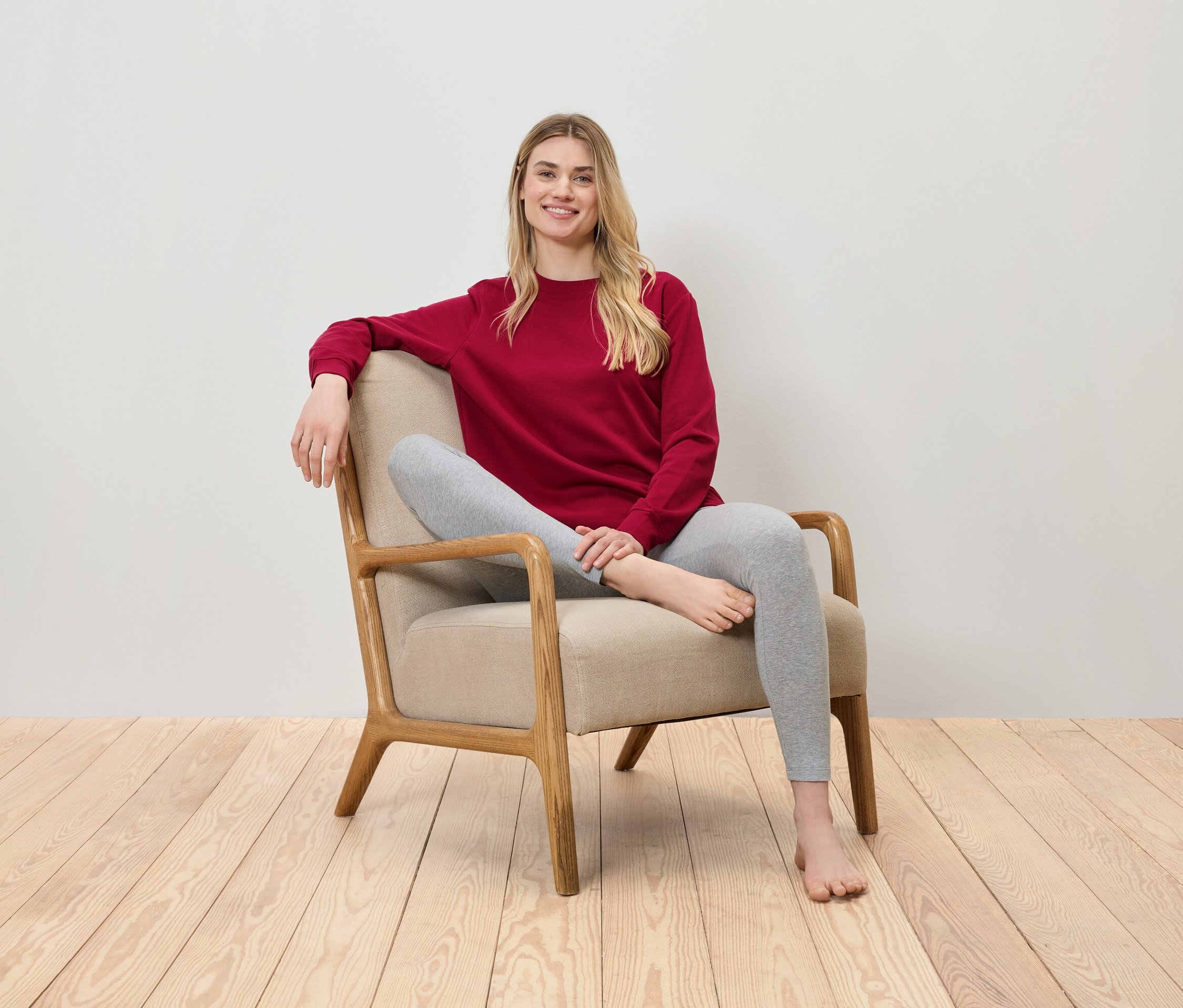 Blonde Frau sitzt in einem beigen Sessel, trägt ein rotes Sweatshirt und graue Leggings.