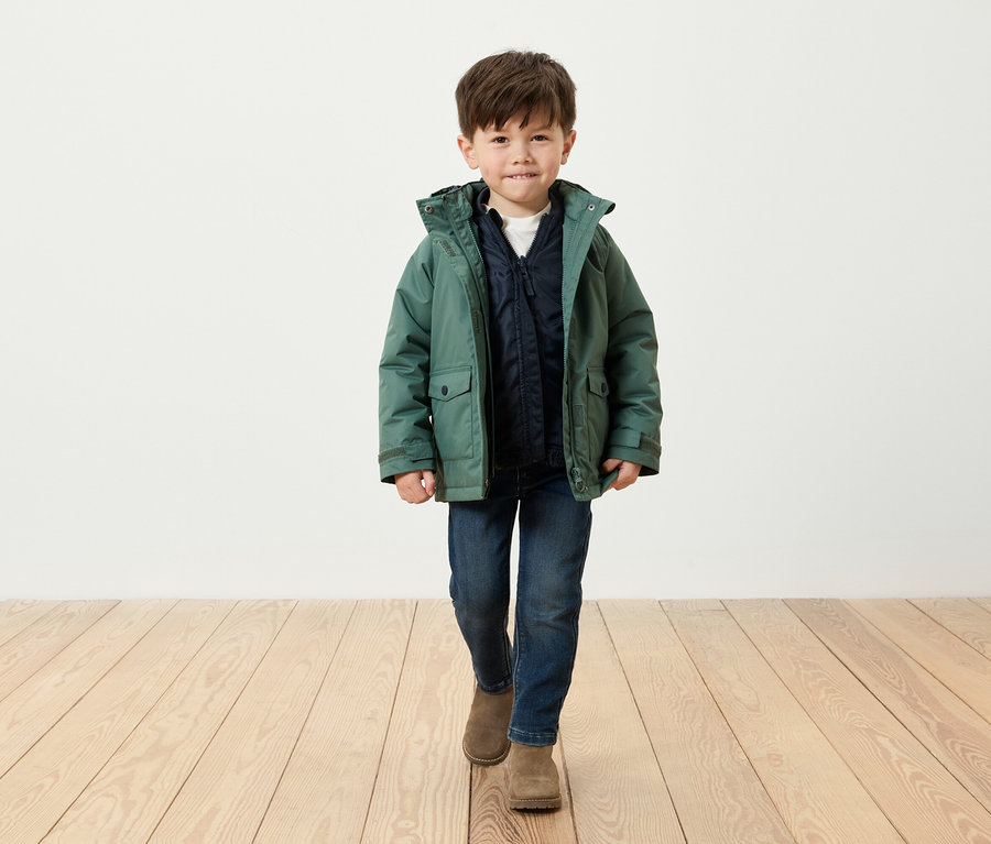 Ein Junge in einer grünen Jacke, blauen Jeans und braunen Stiefeln geht auf einem Holzboden.