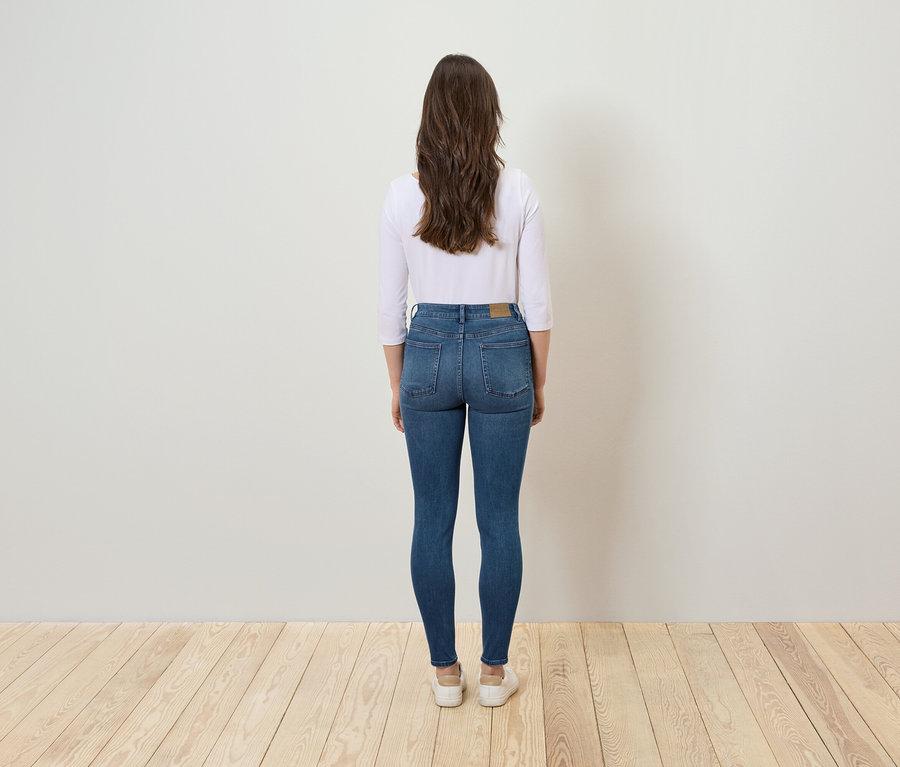 Frau steht mit dem Rücken zur Kamera, trägt blaue Jeans und eine weiße Bluse.