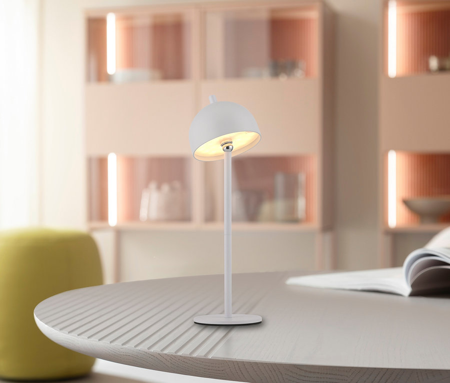 Eine weiße Lampe steht auf einem grauen, runden Tisch. Im Hintergrund befinden sich ein gelber Hocker und Schränke.