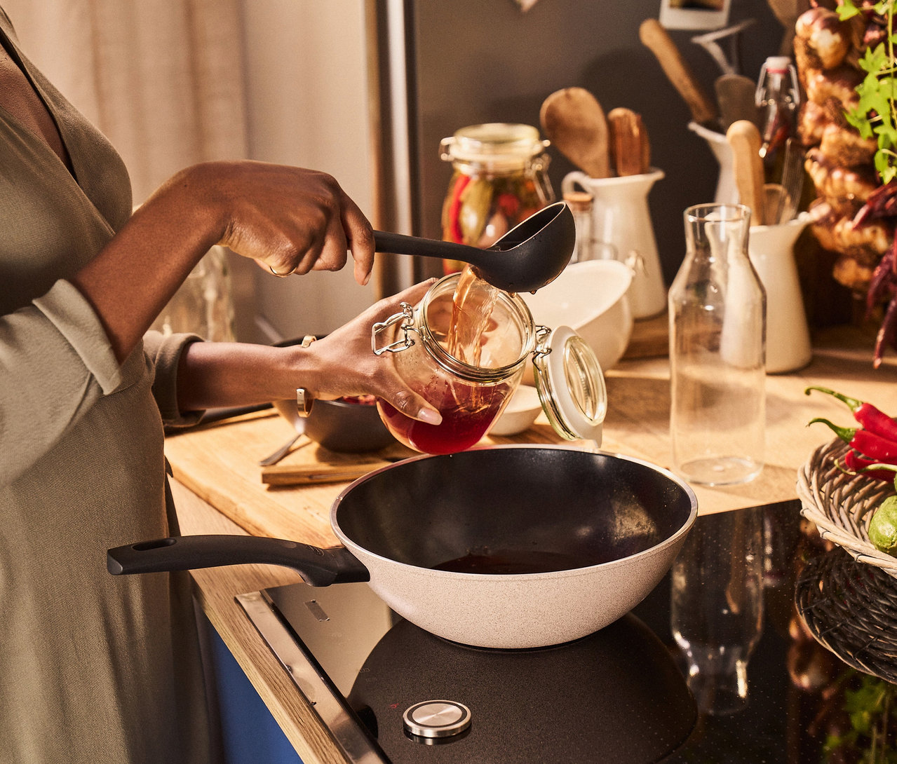 Frau gießt mit einer Schöpfkelle Flüssigkeit aus einem Glas in einen BALLARINI Ledro Wok (28 cm).