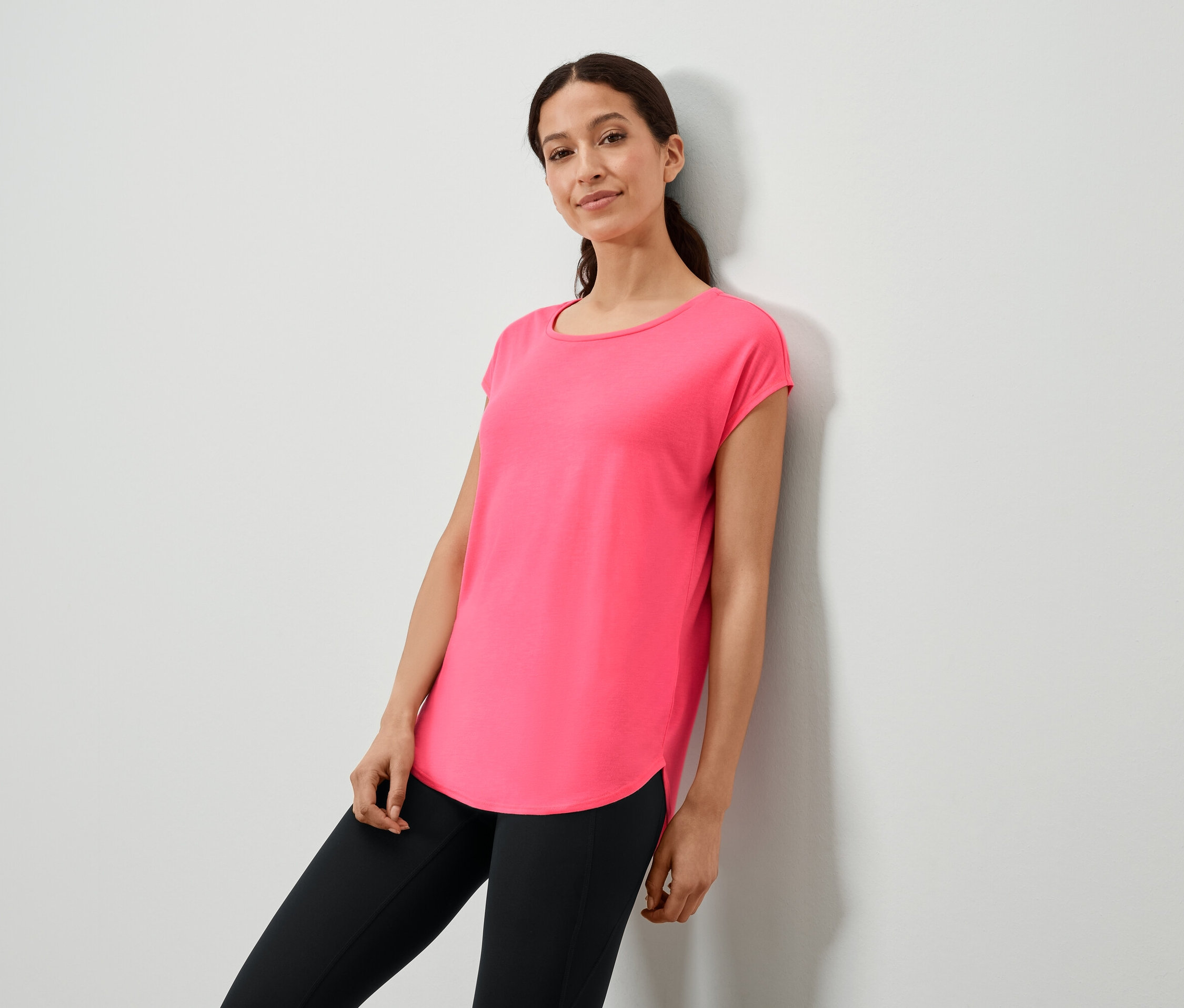 Frau lehnt an einer Wand und trägt ein pinkes T-Shirt und schwarze Leggings.