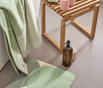 Grüne Komfort-Badematte, XL-Badetuch mit organischen Formen und Halter mit Seifenspender neben der Badewanne. Auf einem Holzhocker liegen 2 LED-Echtwachskerzen und ein offenes Buch.