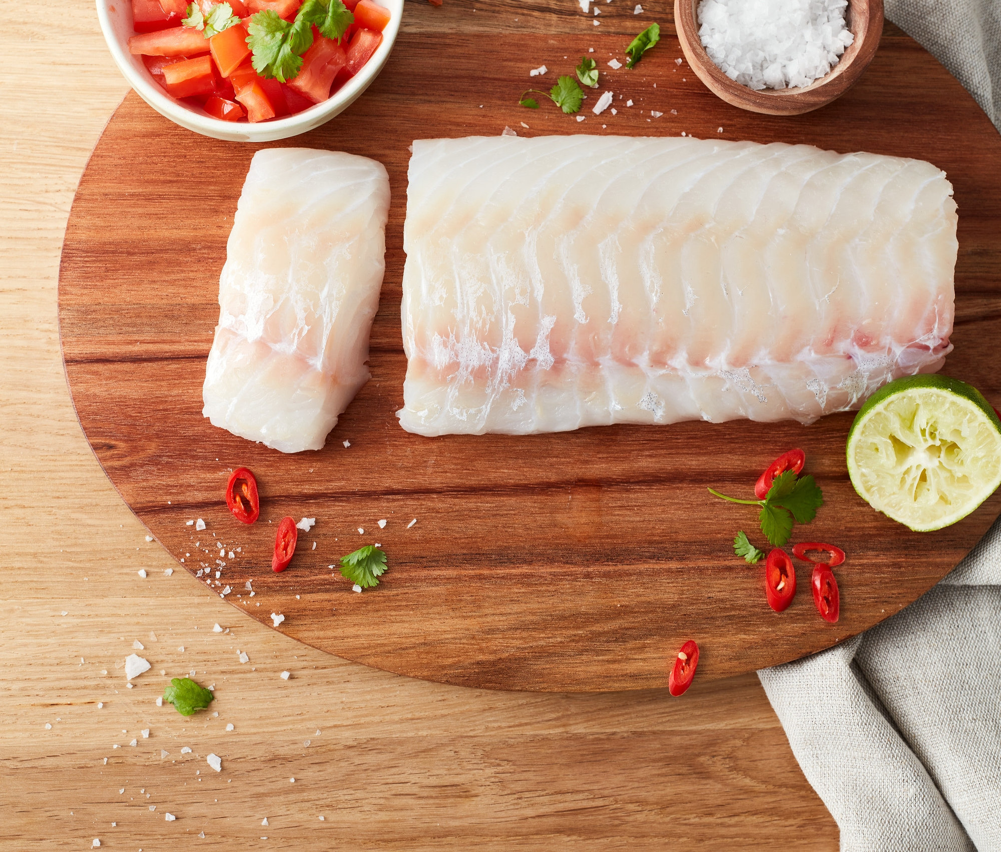Zwei Stücke Kabeljau liegen auf einem Holzbrett mit geschnittenen Tomaten, Salz, Chili und Limette.