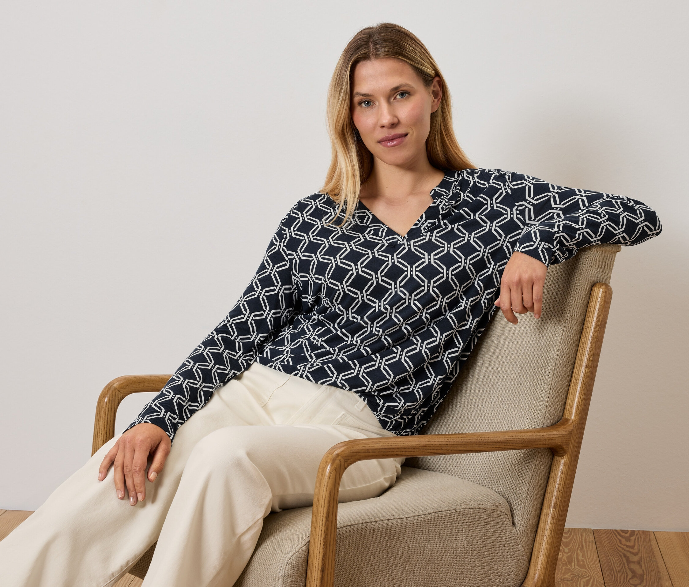 Frau sitzt in einem Sessel und trägt ein blau-weißes Oberteil und cremefarbene Hosen.